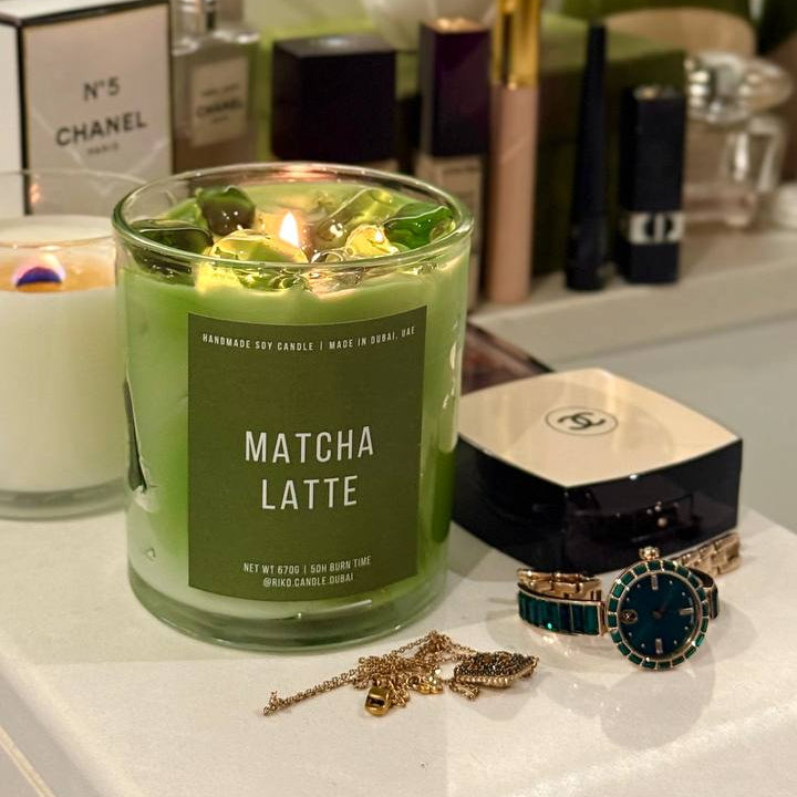 Green 'Matcha Latte' candle on a surface with various items including perfume bottles and jewelry.